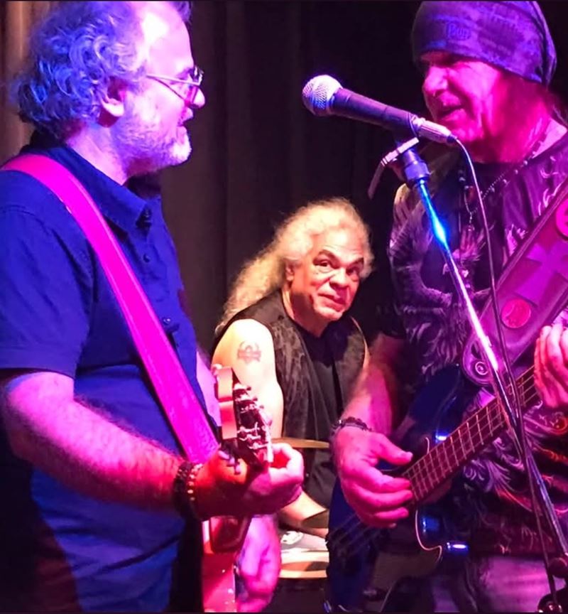 Guitarists sharing a microphone under club lights, with the drummer behind